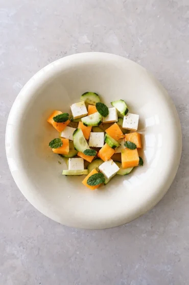 Cantaloupe cucumber vegan feta salad with olive oil, fresh mint, and black pepper in a beige bowl on a soft stone background.