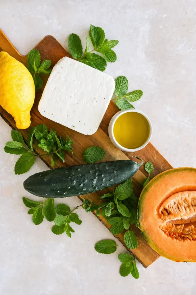 Ingredients for cantaloupe salad arranged on a wooden board, including half a cantaloupe, cucumber, lemon, fresh mint, olive oil, and a block of vegan feta.