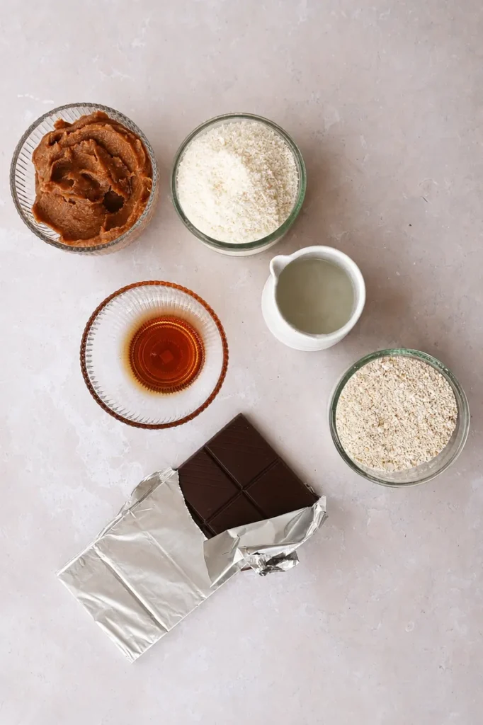Easy Vegan Twix Bars with Dates ingredients arranged on a neutral stone surface, including dates, oats, coconut, maple syrup, chocolate, and plant milk.