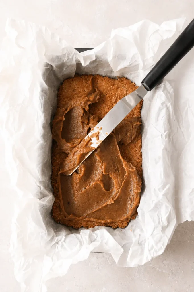 Easy Vegan Twix Bars with Dates caramel layer being spread over the baked base with an offset spatula.
