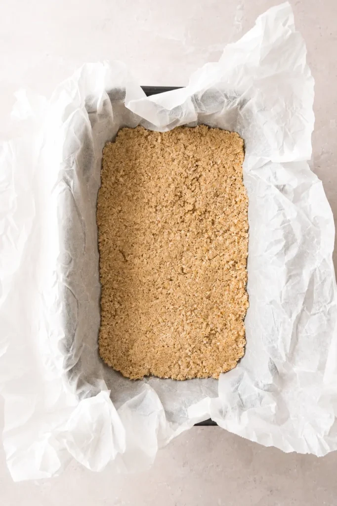 Easy Vegan Twix Bars with Dates crumb mixture being transferred onto parchment paper with a spoon before shaping the base.