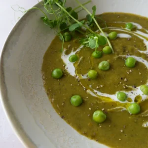 Split pea soup recipe close-up with half of a rustic bowl showing creamy green soup, a swirl of cream, green peas, cracked pepper, and fresh pea shoots.