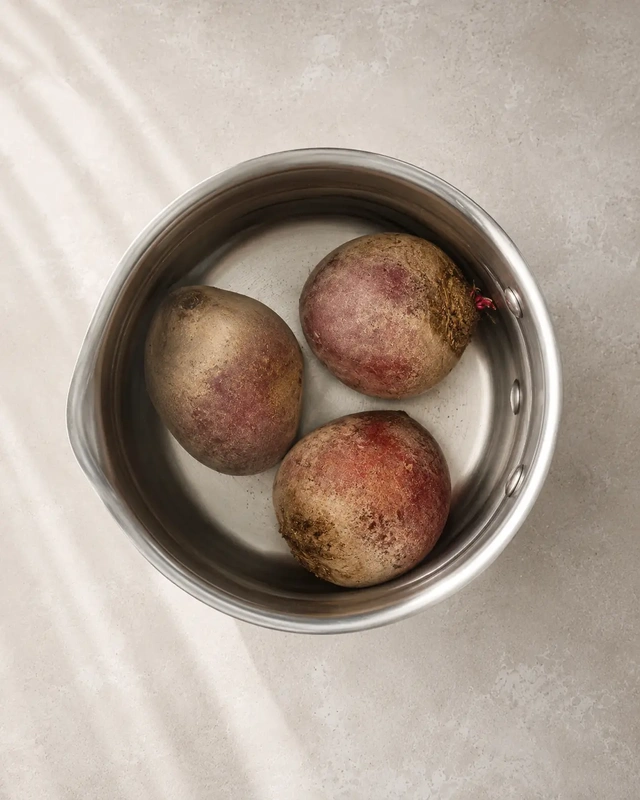 Whole beetroot in a stainless steel pot filled with water, photographed from above on a light stone surface in a minimal editorial style for a how to boil beetroot guide.