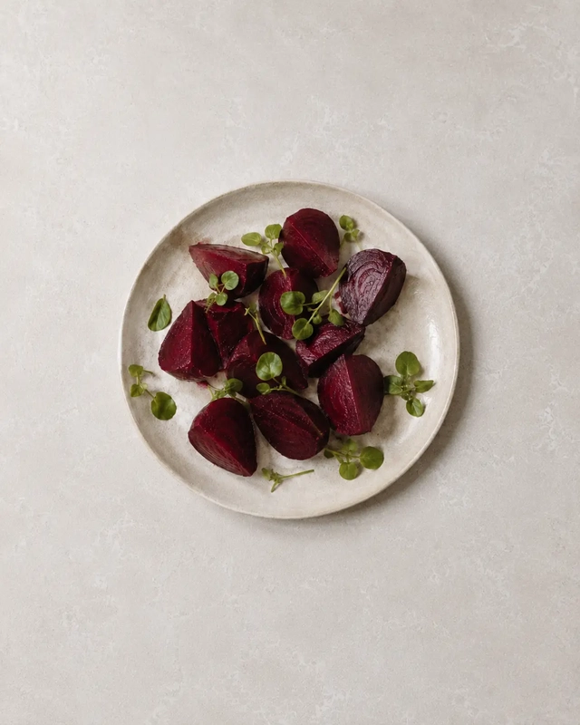 Boiled beetroot wedges with watercress on a ceramic plate, arranged on a neutral stone surface in a minimal editorial food style.