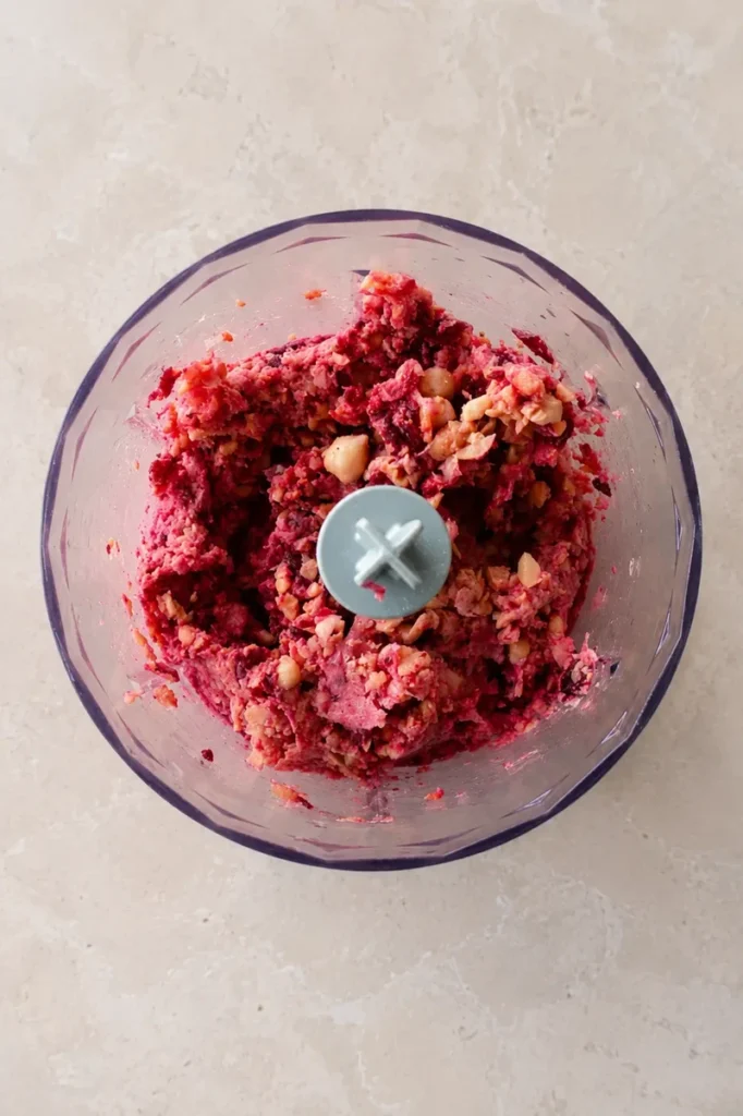 Pink hummus partially blended in a food processor, showing chopped beetroot and chickpeas before reaching a smooth texture.