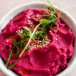 Pink hummus in a white bowl, topped with olive oil, sesame seeds, and fresh greens, shown in a close-up view.