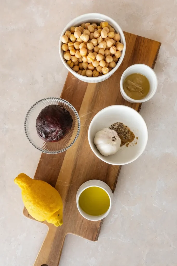 Pink hummus ingredients arranged on a wooden board, with chickpeas, beetroot, lemon, garlic, olive oil, tahini, and spices ready for preparation.
