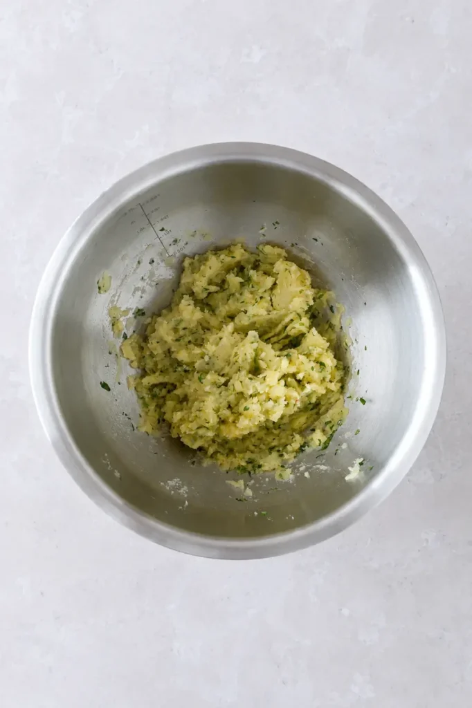 Potato pure mashed mixture in a stainless steel bowl, with potatoes combined with chopped herbs in an overhead view.