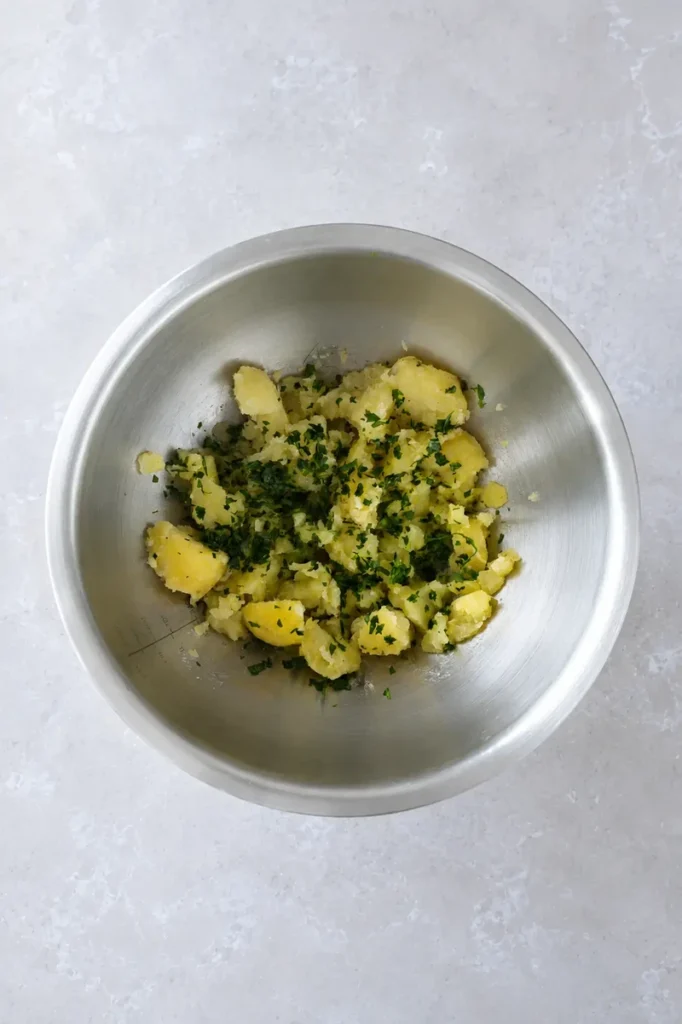 Potato puree partially mashed potatoes in a stainless steel bowl, mixed with fresh chopped parsley before becoming smooth.