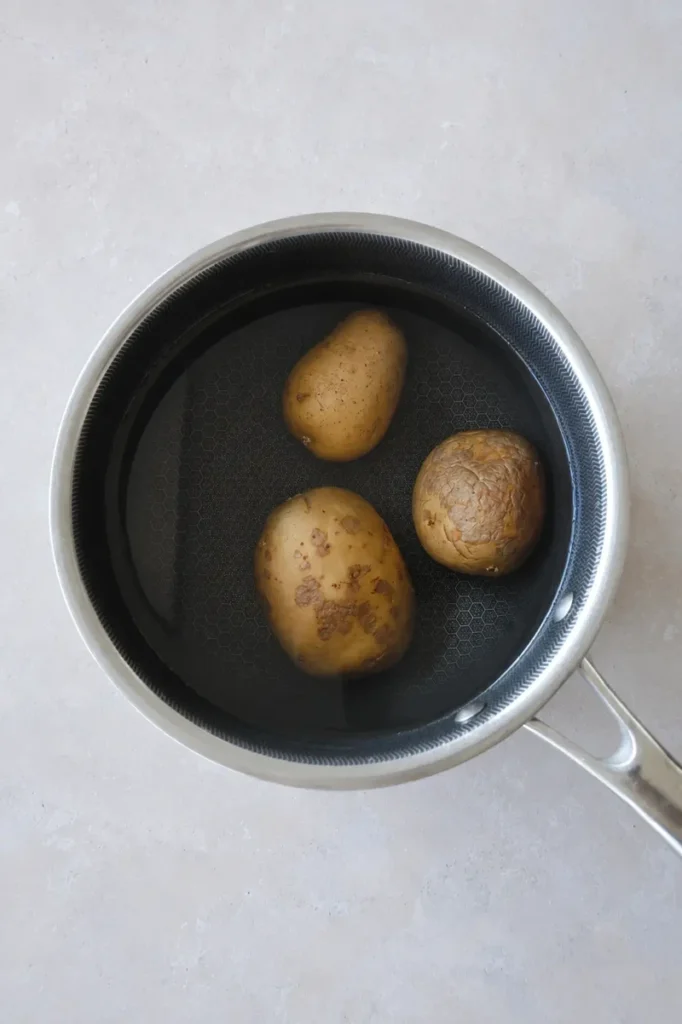 Potato puree potatoes in a stainless steel pan filled with water, shown from above before cooking on a light neutral surface.