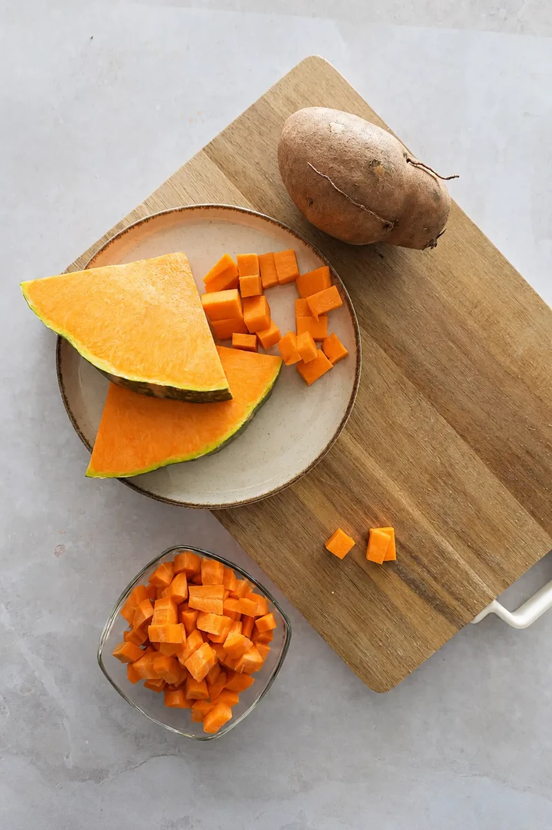 Pumpkin and red lentil soup prep with sliced pumpkin, diced pumpkin, and sweet potato on a wooden board.