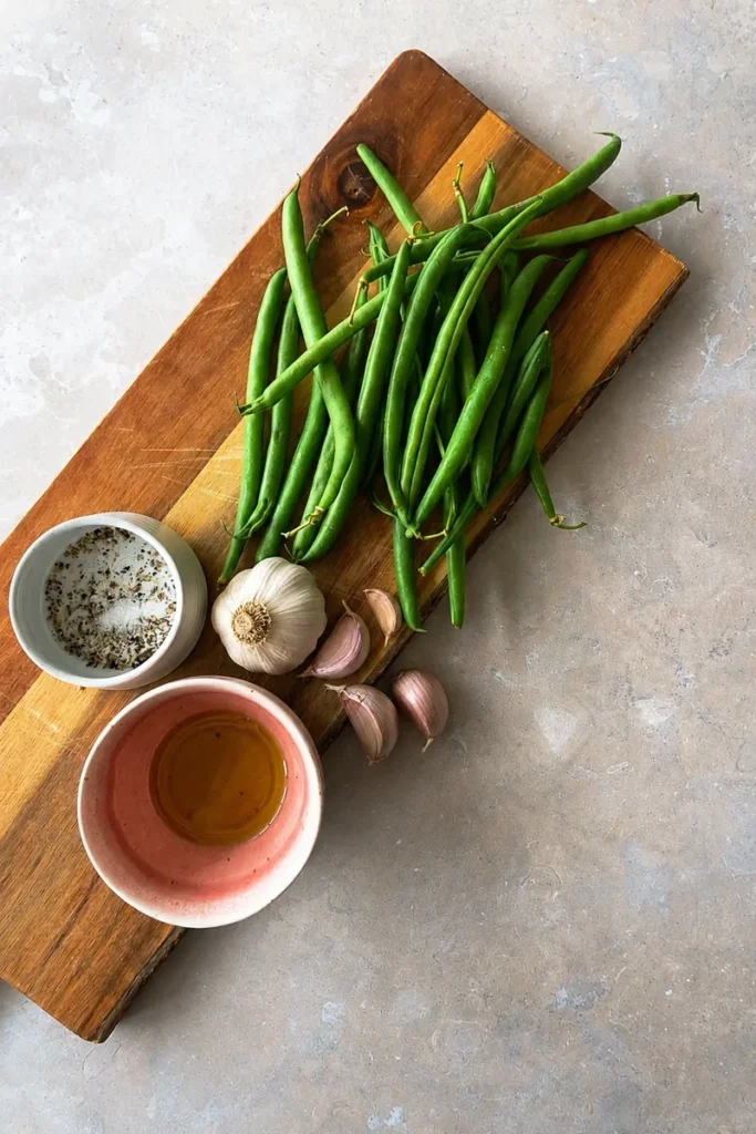 Roasted green beans ingredients on a wooden cutting board, including fresh green beans, olive oil, salt, and black pepper.