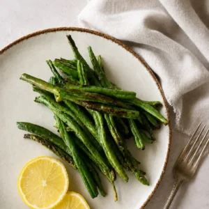 Roasted green beans recipe served on a rustic ceramic plate with lemon slices, photographed from above on a light stone background.