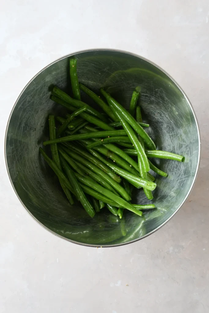 Roasted green beans recipe prep with fresh green beans in a stainless steel mixing bowl on a light stone surface.