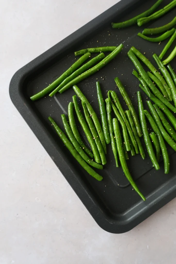 Roasted green beans arranged on a black oven tray with olive oil, garlic, salt, and pepper before baking.