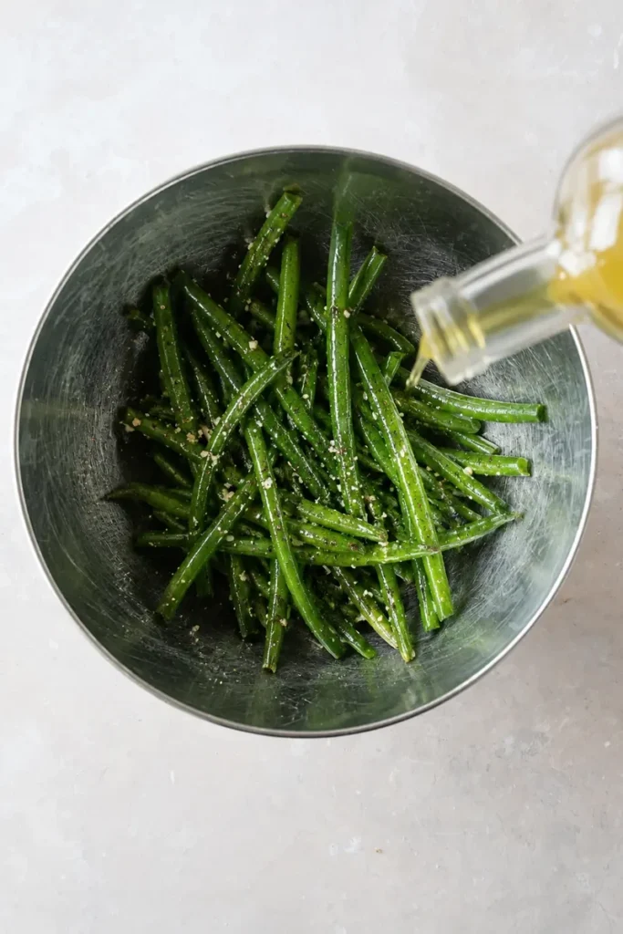 Roasted green beans recipe seasoning step with olive oil poured over green beans, salt, pepper, and garlic in a stainless steel bowl.