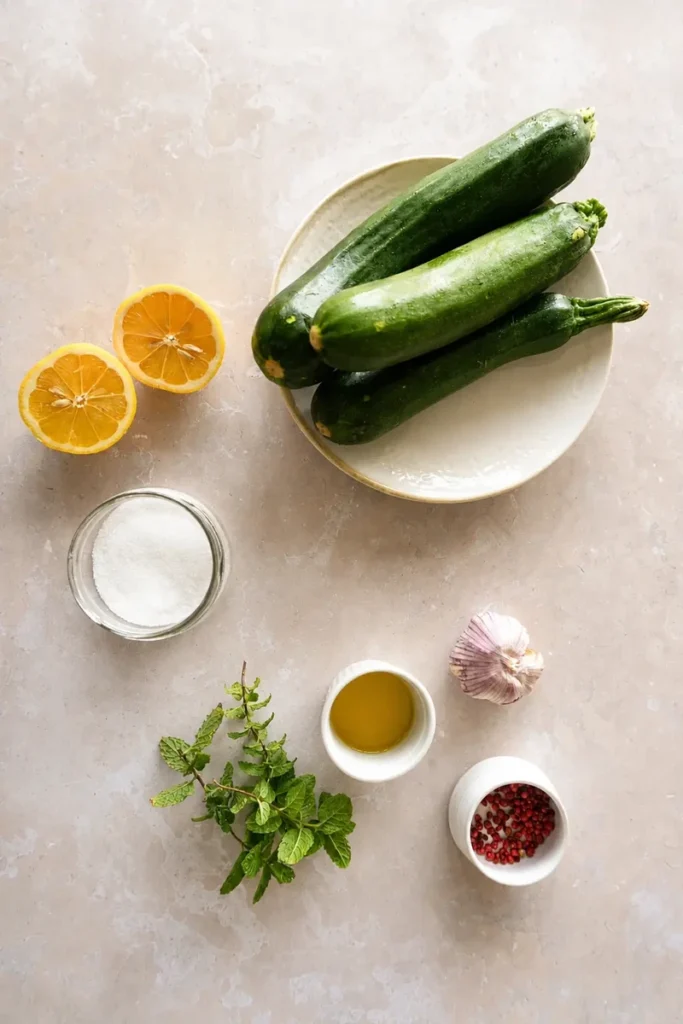 Zucchini recipe ingredients with fresh zucchini, lemon, garlic, olive oil, mint, salt, and pink peppercorns arranged on a light stone surface.