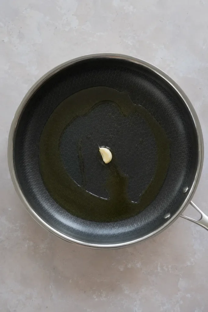 Zucchini recipe step with olive oil and a single garlic clove in a nonstick pan, photographed from above before sautéing.