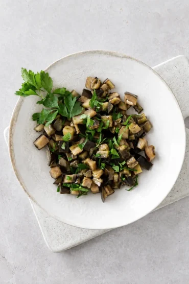 Sauteed eggplants with garlic and herbs served in a ceramic bowl with fresh parsley on top.