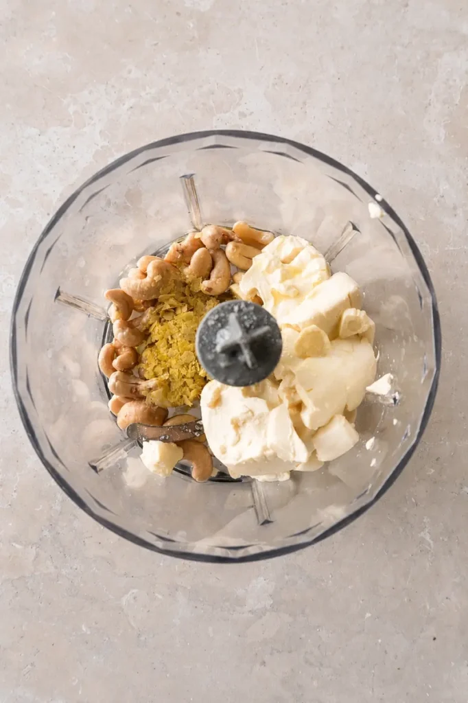 Vegan aioli ingredients in a food processor with soaked cashews, vegan cream cheese, nutritional yeast, garlic, and olive oil.