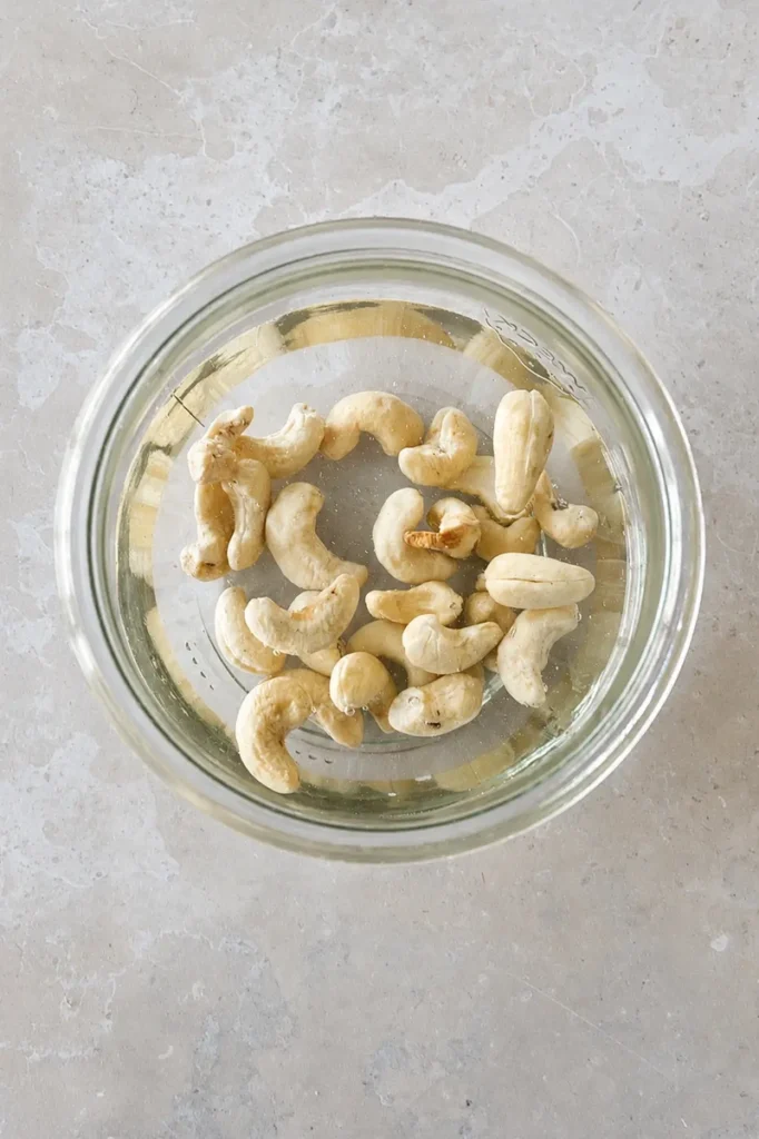 Vegan aioli cashews soaking in water in a glass jar on a neutral kitchen surface.
