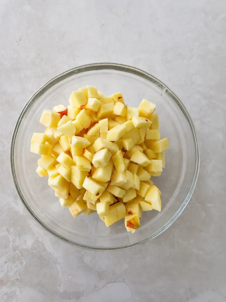 Vegan apple cake recipe ingredients with diced apples in a glass bowl on a light stone background.