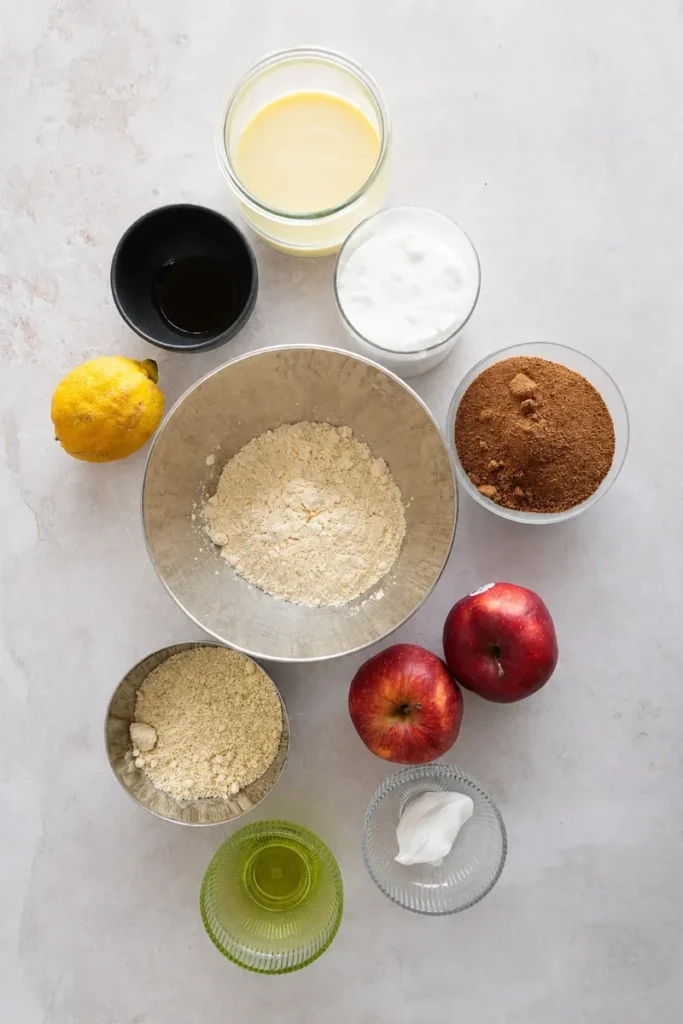 Vegan apple cake ingredients arranged on a light stone surface with apples, flour, sugar, lemon, plant-based milk, oil, and yogurt.