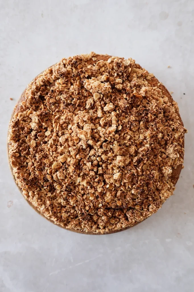 Vegan apple cake topped with golden oat crumble, photographed from above in a round cake pan on a light neutral background.