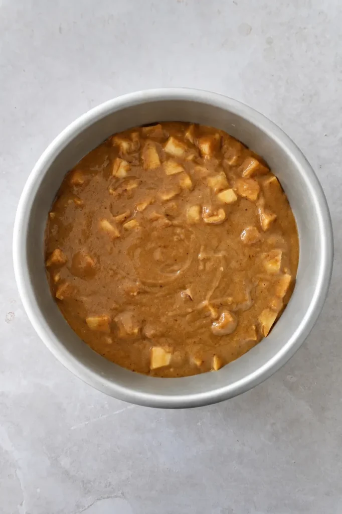 Vegan apple cake batter with apple pieces poured into a round cake pan, photographed from above before baking.