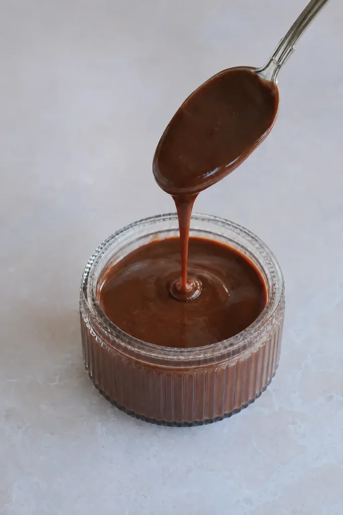 chocolate sauce in a small glass jar with a silver spoon lifting a thick stream of glossy chocolate sauce, photographed on a light neutral background in a minimal food styling scene