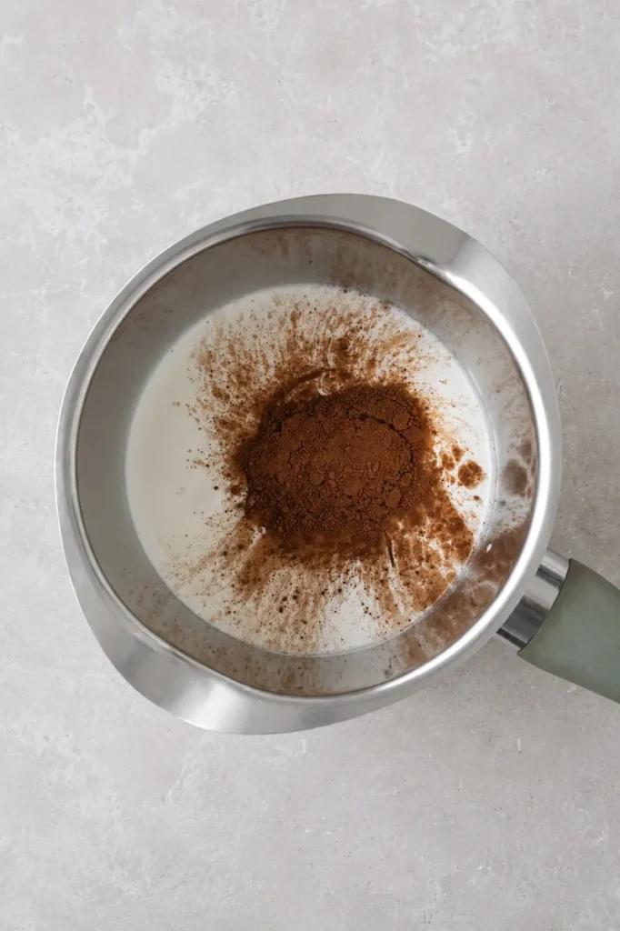 Vegan chocolate sauce preparation with cocoa powder added to plant-based milk in a stainless steel saucepan.
