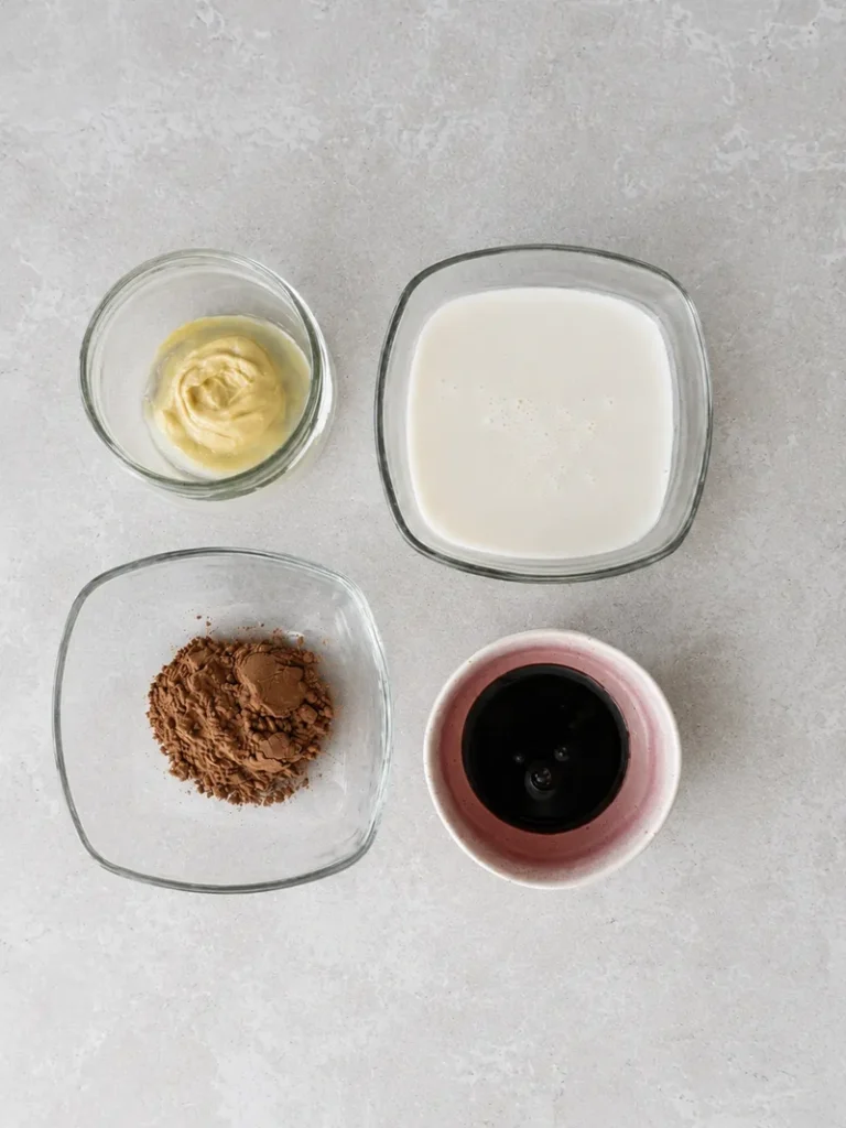 Vegan chocolate sauce ingredients arranged on a neutral countertop with cocoa powder, plant-based milk, maple syrup, and creamy nut butter.