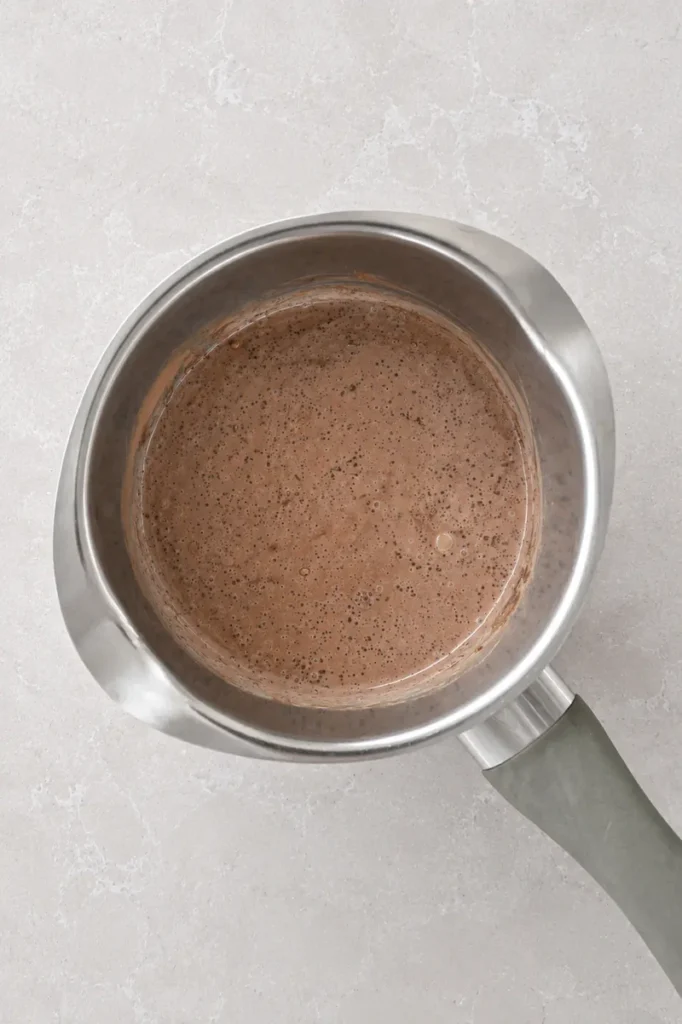 Vegan chocolate sauce mixture stirred in a saucepan until smooth, creamy, and lightly speckled with cocoa.