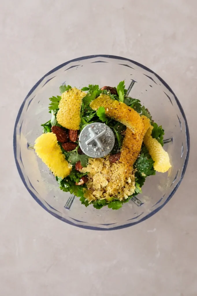 Vegan pesto pasta preparation with parsley, almonds, lemon zest, and olive oil inside a food processor.