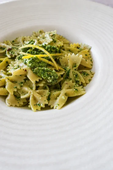 Vegan pesto pasta with bow-tie pasta, creamy green pesto, and lemon zest served in a wide ceramic bowl.