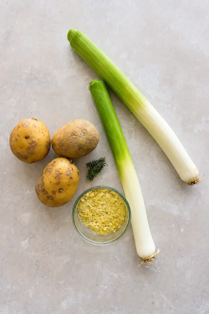 Vegan potato leek soup ingredients with potatoes, leeks, thyme, and nutritional yeast arranged on a light stone surface.