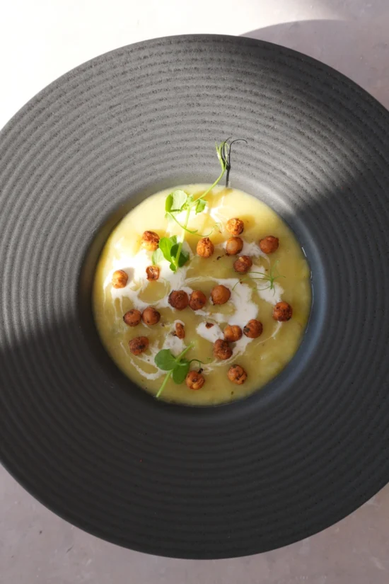 Vegan potato leek soup served in a black bowl, topped with crispy chickpeas, a swirl of cream, and fresh greens in an overhead view.