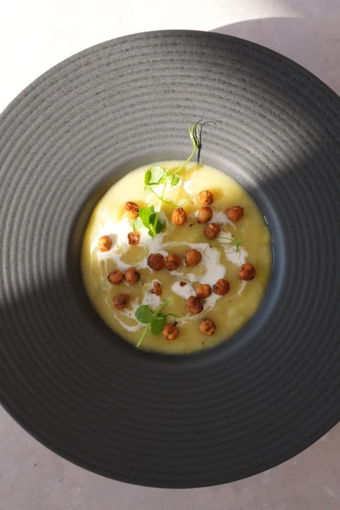 Vegan potato leek soup served in a black bowl, topped with crispy chickpeas, a swirl of cream, and fresh greens in an overhead view.