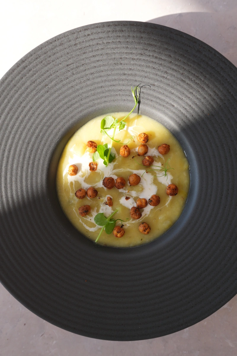 Vegan potato leek soup served in a black bowl, topped with crispy chickpeas, a swirl of cream, and fresh greens in an overhead view.