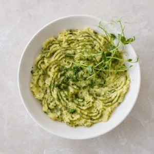 Potato puree served in a white bowl, topped with chopped parsley, olive oil, and fresh greens in an overhead view.