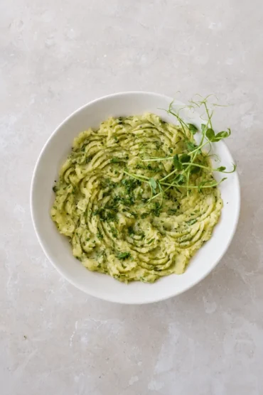 Potato puree served in a white bowl, topped with chopped parsley, olive oil, and fresh greens in an overhead view.