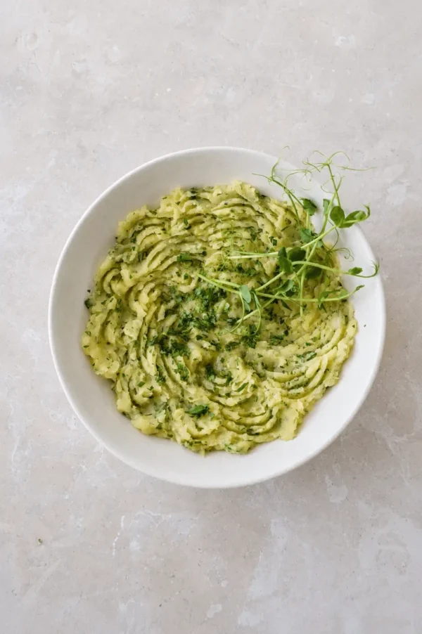 Potato puree served in a white bowl, topped with chopped parsley, olive oil, and fresh greens in an overhead view.