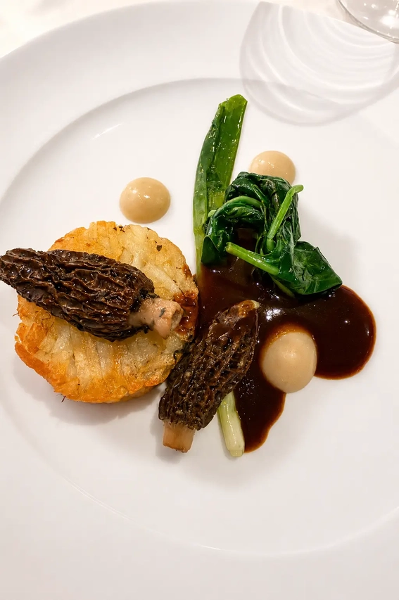 Gauthier Soho vegan fine dining dish with morel mushrooms, crispy potato, wilted greens, and glossy dark sauce on a white plate.
