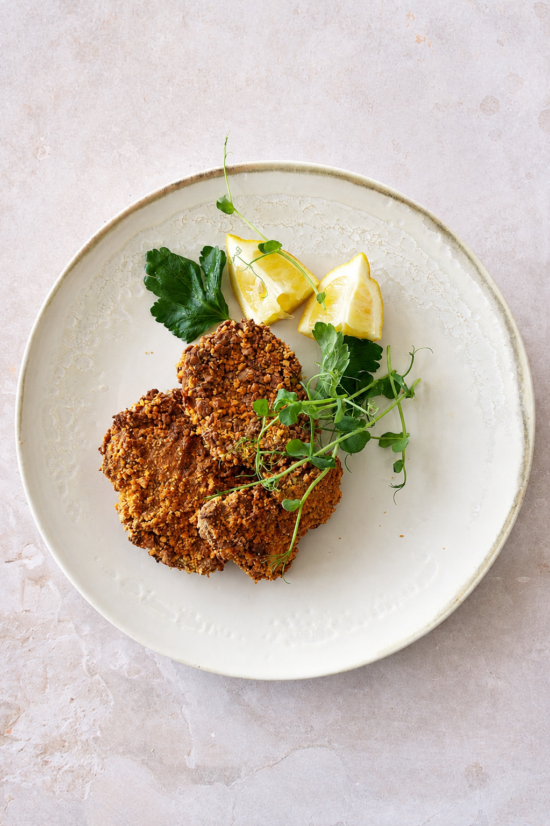 Vegan schnitzel served with lemon wedges, fresh parsley, and pea shoots on a pale ceramic plate.
