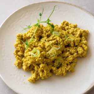 vegan tofu scramble on white plate, close-up of golden tofu scramble served on a white ceramic plate, topped with green onion rings and pea shoots on a soft neutral background.