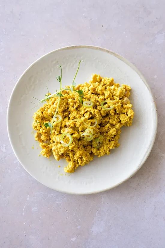 vegan tofu scramble on white plate, top-down view of golden tofu scramble plated on a white ceramic dish, garnished with green onion slices and pea shoots on a light neutral surface.