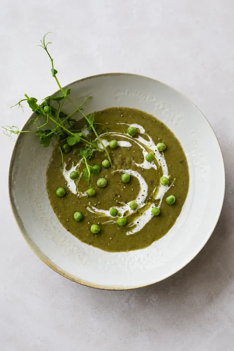vegan Split pea soup recipe served in a rustic ceramic bowl with a swirl of cream, green peas, cracked pepper, and fresh pea shoots on a light stone background.