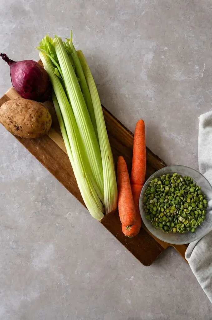 Split pea soup recipe ingredients with green split peas, celery, carrots, potato, and red onion arranged on a wooden board.