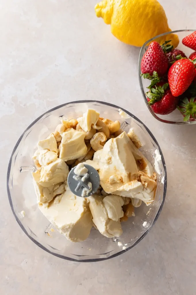Vegan strawberry cheesecake filling ingredients in a food processor with cashews, vegan cream cheese, and lemon nearby.