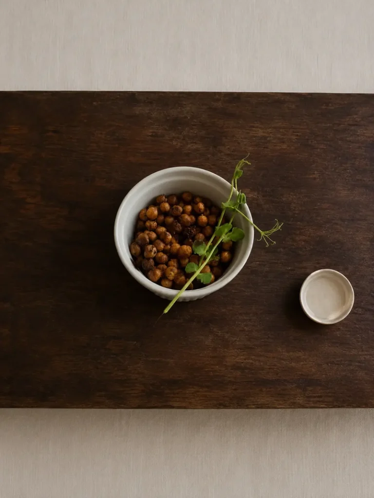 Umami foods Overhead still life of a white ceramic bowl filled with roasted chickpeas and topped with delicate pea shoots, placed on a dark rustic wooden table beside a small neutral-toned dish, in a warm minimalist editorial style.